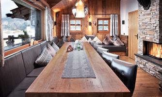 Cozy dining area with panoramic views and fireplace. The spacious solid wood table accommodates sociable gatherings in a stylish atmosphere. Soft cushions, warm wooden walls, and decorative curtains emphasize the cozy chalet flair. The large panoramic window opens up views of the mountain landscape, while the open fireplace creates a particularly atmospheric setting – ideal for long evenings with family or friends.