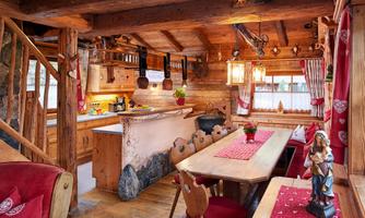 Cozy cabin atmosphere with a large dining table and open kitchen. This lovingly furnished living room in rustic cabin style invites you to linger. The spacious solid wood dining table offers plenty of space for sociable gatherings and family meals. The open kitchen is designed with natural wood elements and harmoniously blends into the warm ambiance. Traditional details such as carved chairs with heart motifs, decorative curtains with alpine hearts, and a rustic wooden ceiling emphasize the charming country house style. A special highlight is the handcrafted decorations and atmospheric lighting that create a cozy ambiance – ideal for unforgettable cabin evenings.