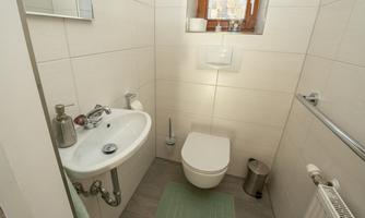 A small, modern bathroom with a toilet and a sink. The walls are light-tiled, and a window provides natural light.