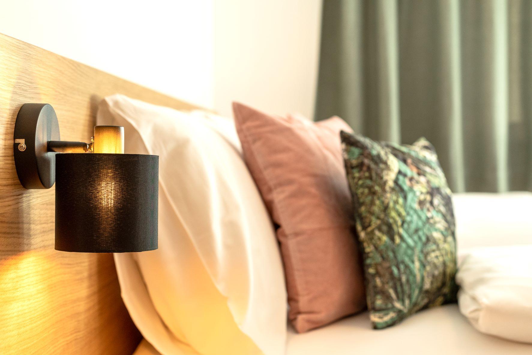 A cozy bedroom with a wooden bed and decorative pillows. A stylish lamp hangs on the wall.