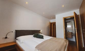A modern bedroom with a large bed and warm wood details. The walls are light and well-lit, creating an inviting atmosphere. A small, private bathroom with a sink and shower is also available.