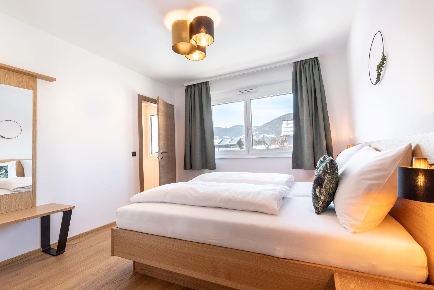 A modern bedroom with a large window and a cozy atmosphere, as well as an adjoining bathroom. The room is equipped with a wooden bed and a mirror.