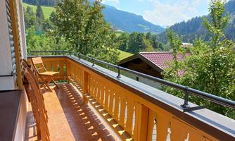 A balcony with wooden chairs and a beautiful view of the mountains. The sky is clear and nature is green and inviting.