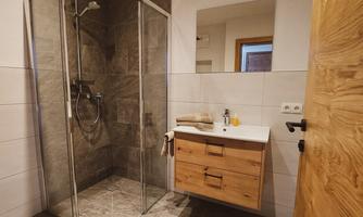 A modern bathroom with a glass shower and a wooden sink. The floor is tiled with gray tiles, and there are towels as well as a bath mat.
