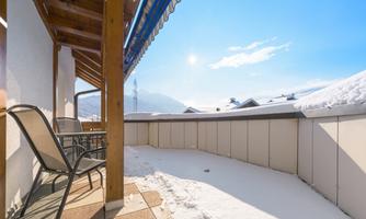 A cozy terrace with a table and chairs, surrounded by snow and a clear blue sky. In the background, snow-covered trees and mountains are visible.