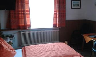 A simple room with a double bed and orange bed covers. In front, there is a table and a chair, and the window lets in a lot of light.