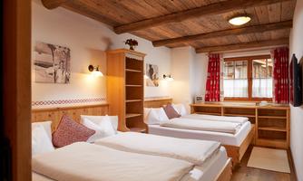 Spacious Quadruple Room – ideal for families or friends<br/>This cozy quadruple room in alpine style offers space and comfort for up to four people. Two generous double beds, lovingly decorated wooden furniture, and traditional fabrics create a warm and inviting atmosphere. The rustic wooden ceiling, the red curtains with heart motifs, and nostalgic black-and-white photographs emphasize the charming cabin character. Ample storage space, a TV, and natural light ensure a pleasant stay – perfect for shared holiday moments with the whole family or good friends.