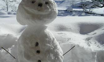 A snowman stands in the foreground, surrounded by snow and a winter landscape. In the background, trees and mountains can be seen under a clear blue sky.