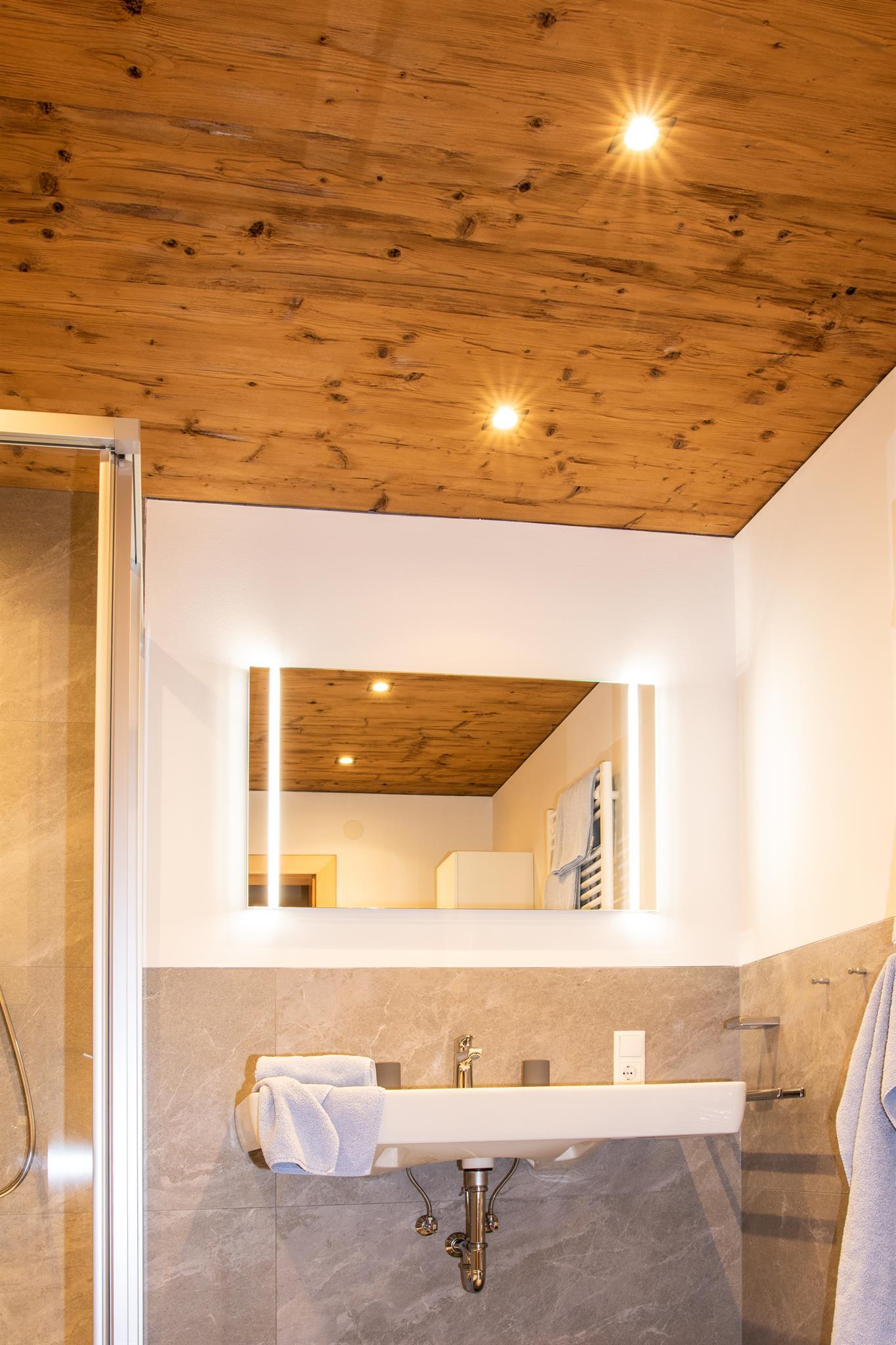 A modern bathroom with a large mirror and a practical shelf. The wooden ceiling and warm lights create an inviting atmosphere.