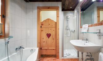 The stylishly decorated bathroom combines modern functionality with cozy Alpine charm. It includes a comfortable bathtub, a modern shower cubicle, as well as a cosmetic mirror and a hairdryer. A large window provides natural daylight and creates a pleasant, inviting atmosphere. This bathroom offers a cozy ambiance for relaxing moments of well-being.