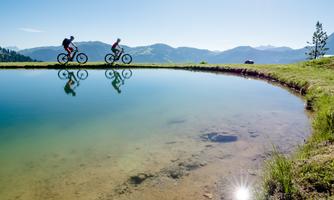Two cyclists ride along a quiet pond in a beautiful mountain landscape. The clear sky and the reflecting mountains create a peaceful atmosphere.