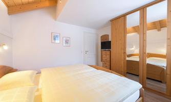 A cozy, comfortably furnished bedroom with a double bed and solid wood furniture. The large window provides daylight in the room, and the wardrobe offers plenty of space for clothing. There is also a television in the bedroom.
