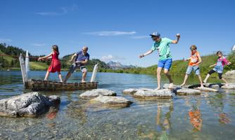 A family is having fun in the water and crossing stones in a clear lake. In the background, green hills and blue sky can be seen.