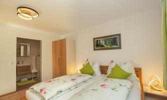 A bright bedroom with two beds, decorated with colorful floral bedding and green pillows. In the background, a modern bathroom is visible.