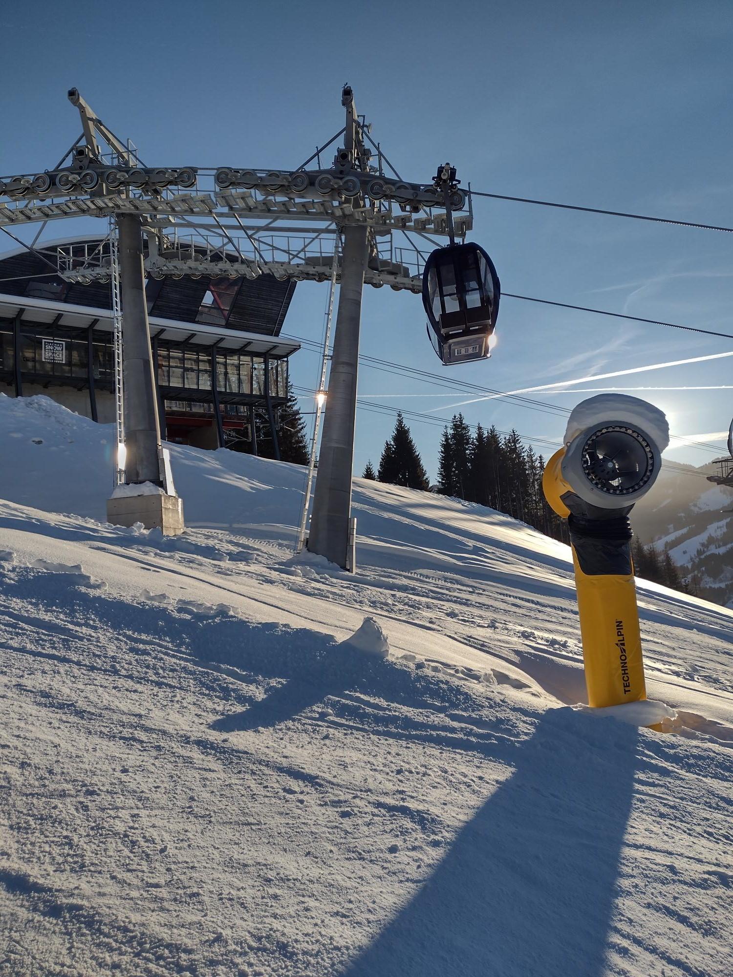 A cable car travels over a snowy landscape. In the foreground, a snow cannon is visible.