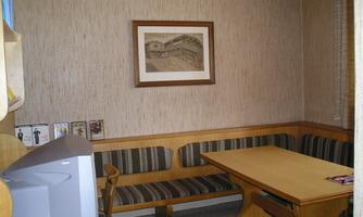 A small room with a wooden bench and a table. A painted picture hangs on the wall, and there is a television in the corner.