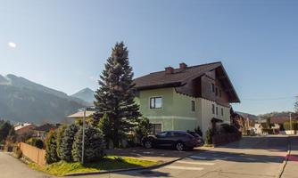 A large house with a light green facade and a black roof. In the foreground, there are tall fir trees and a clear mountain landscape in the background.