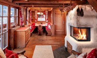 Rustic living room with fireplace and traditional corner bench. Cozy living room in traditional Alpine style with modern comfort. The comfortable tiled stove provides cozy warmth and invites relaxation. The large, surrounding corner bench with soft cushions and heart-decorated pillows offers a perfect spot for shared meals or sociable hours with family and friends. Rustic wooden beams, natural materials, and loving details like embroidered curtains, wood carvings, and decorative elements create an authentic and inviting atmosphere – ideal for unforgettable cabin moments with panoramic views into the valley.
