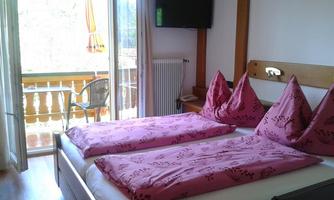 A cozy bedroom with a double bed and pink pillows. The room has a balcony with a view and a television on the wall.