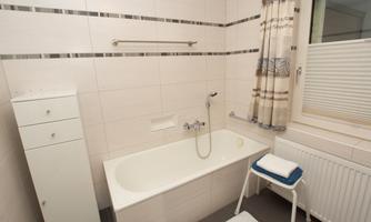 A bright bathroom with a bathtub and a white shelf. In front of the bathtub stands a stool with a towel.