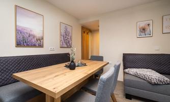 A modern dining room with a wooden table, chairs, and a cozy bench. Decorative pictures of flowers hang on the walls, and there is also a sofa bed in the living-sleeping area.