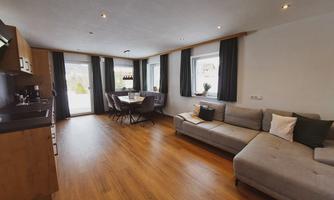 A modern designed living room with a large, comfortable sofa and a dining table for six people. The windows are fitted with dark curtains and offer a view of the outdoor area.