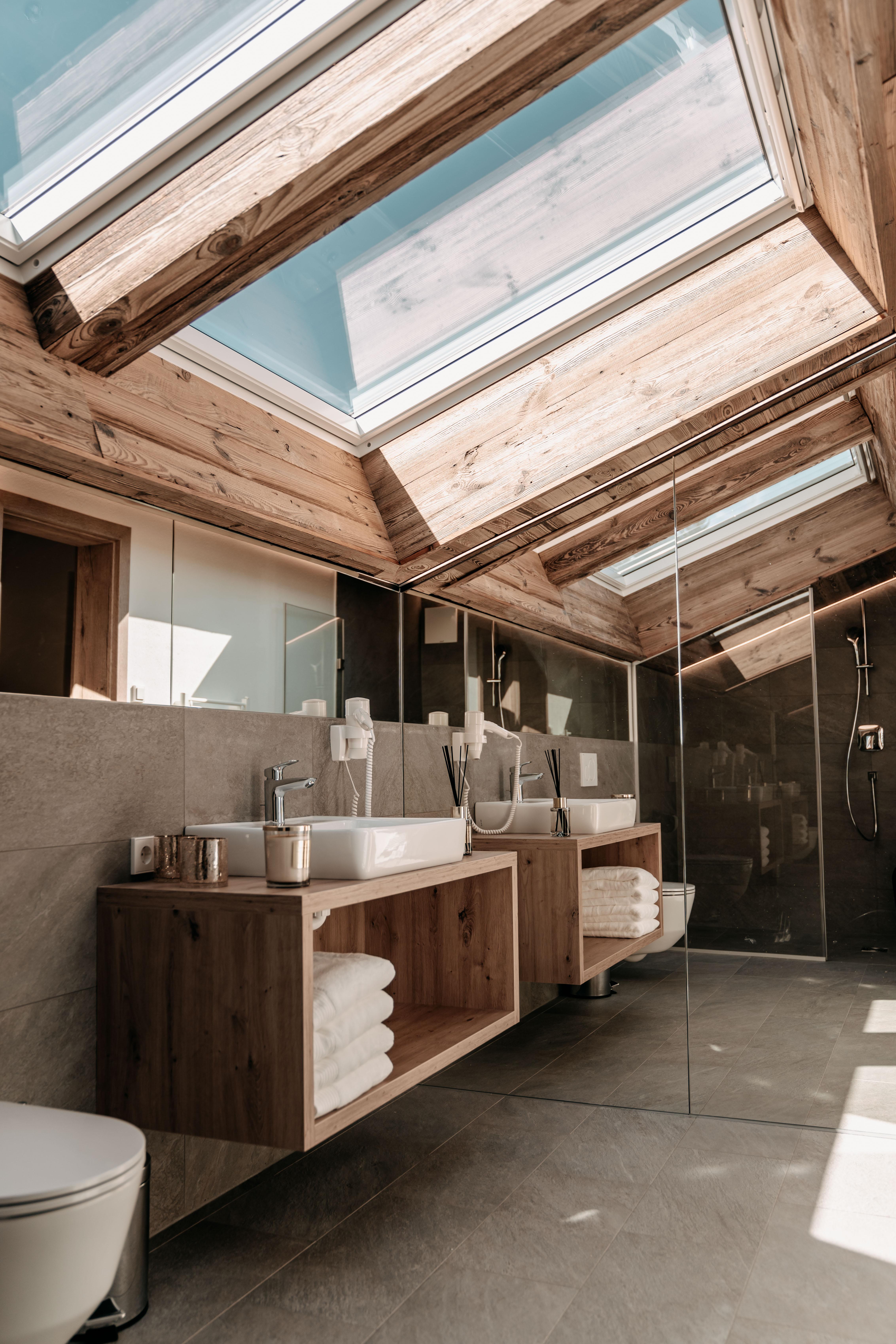 A modern bathroom with a wooden ceiling and large skylights. The space is bright and minimally designed, featuring a sink and towels on a wooden shelf.