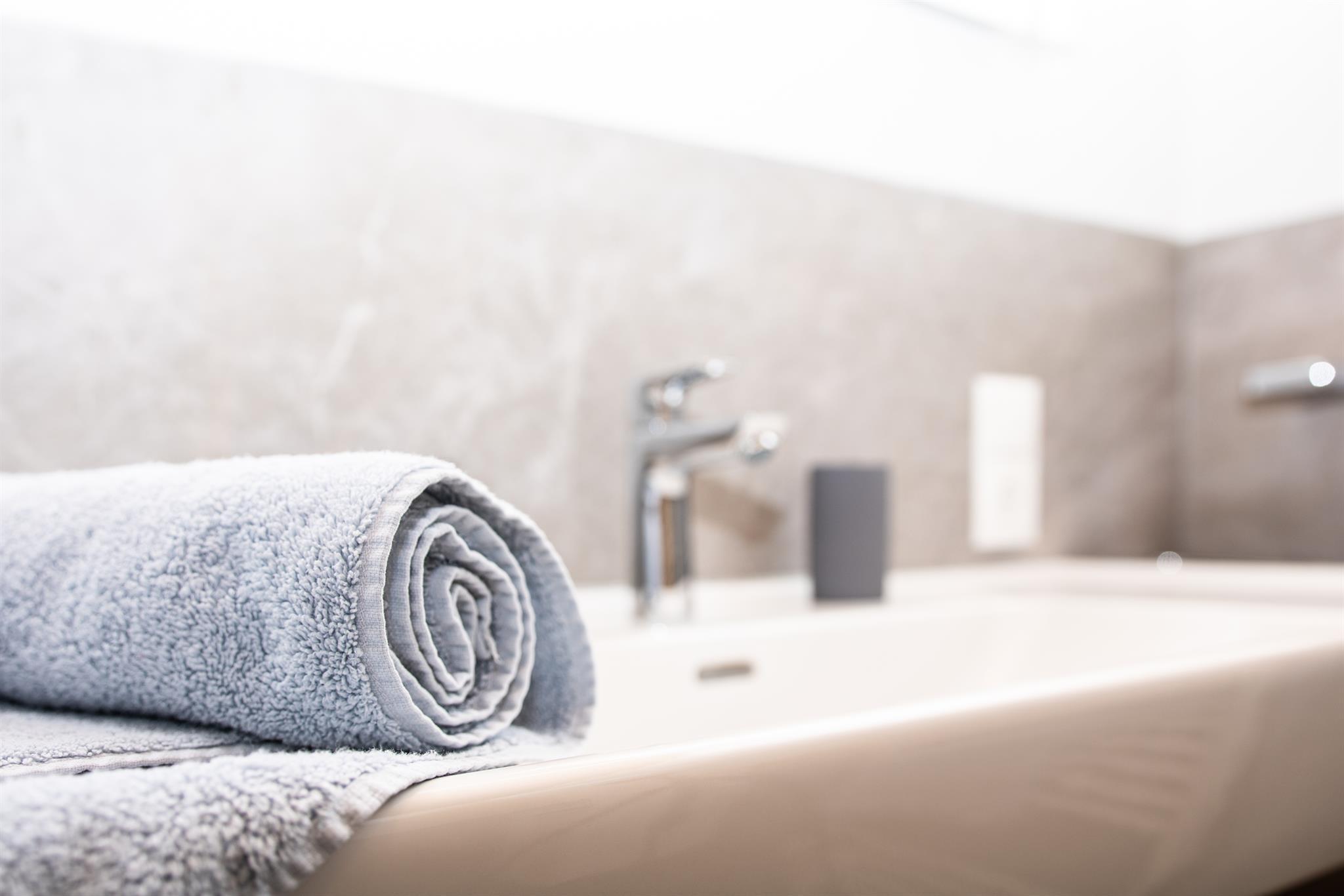 A freshly rolled towel lies on the edge of a modern sink. In the background, elegant faucets and a bright, minimalist wall design can be seen.