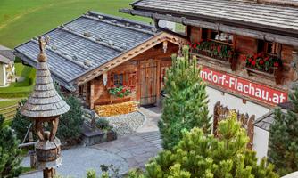 Inviting cabin access in the Alpine village of Flachau. The lovingly designed path to the chalets exudes alpine charm and heartfelt hospitality at first glance. Natural stone, wooden shingles, traditional shutters, and blooming flower boxes shape the authentic appearance. The well-maintained outdoor area harmoniously blends into the surrounding mountain landscape. The large sign reveals upon arrival: Welcome to the Almdorf Flachau – your vacation destination with heart.