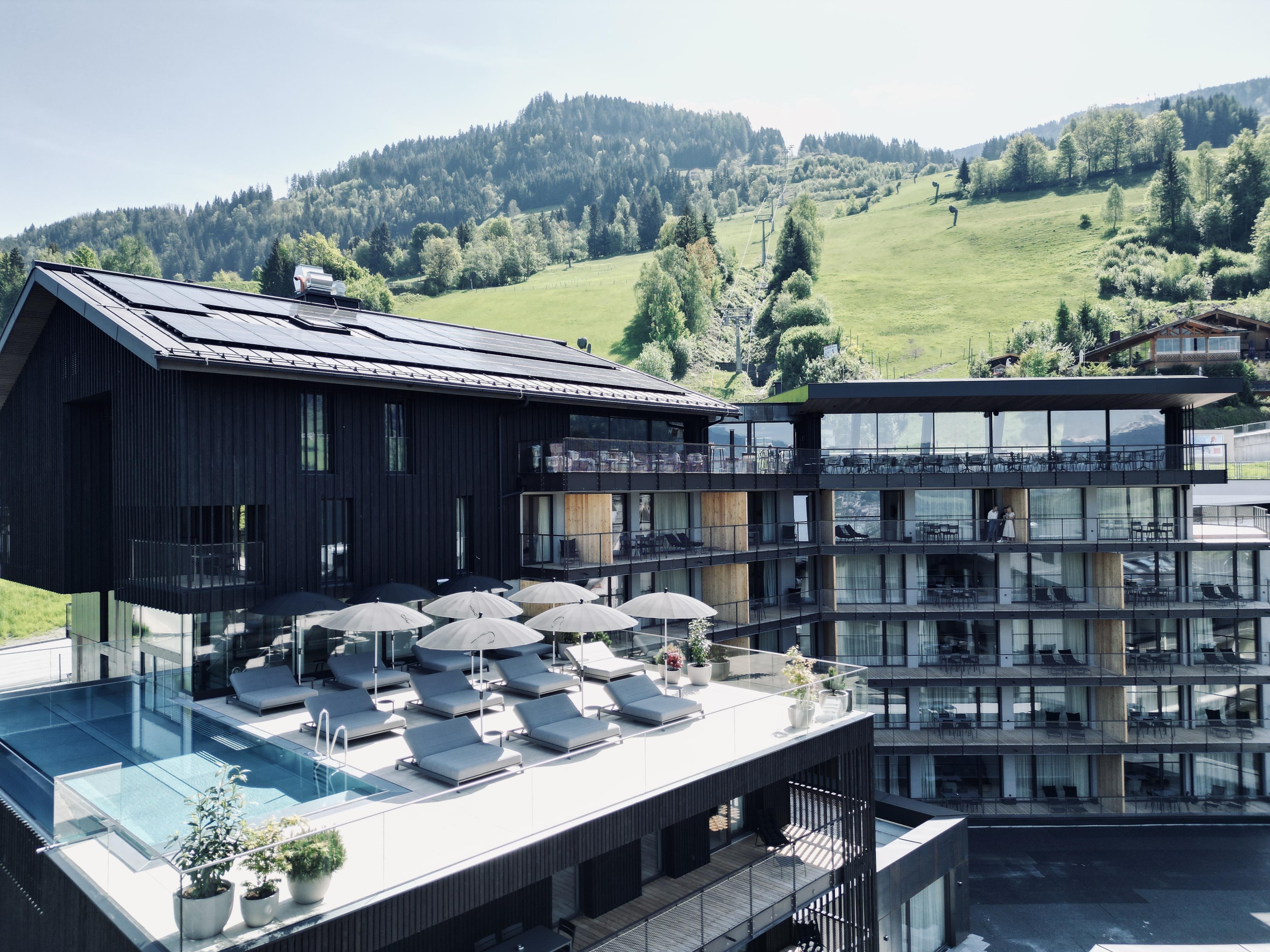 A modern hotel building with large windows and a flat roof. The terrace features lounge chairs and umbrellas with a view of green hills.