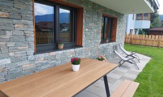 A modern house with stone cladding and large windows. Outside there is a wooden table and two loungers on a well-kept lawn area.