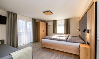 A modern bedroom with a large bed and wooden furniture. The large windows provide plenty of daylight and a bright, inviting atmosphere. The bedroom features a double sofa bed and a television.