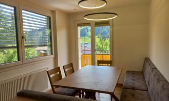 A bright dining area with a large wooden table and a cozy corner bench. Large windows offer a view of the greenery.