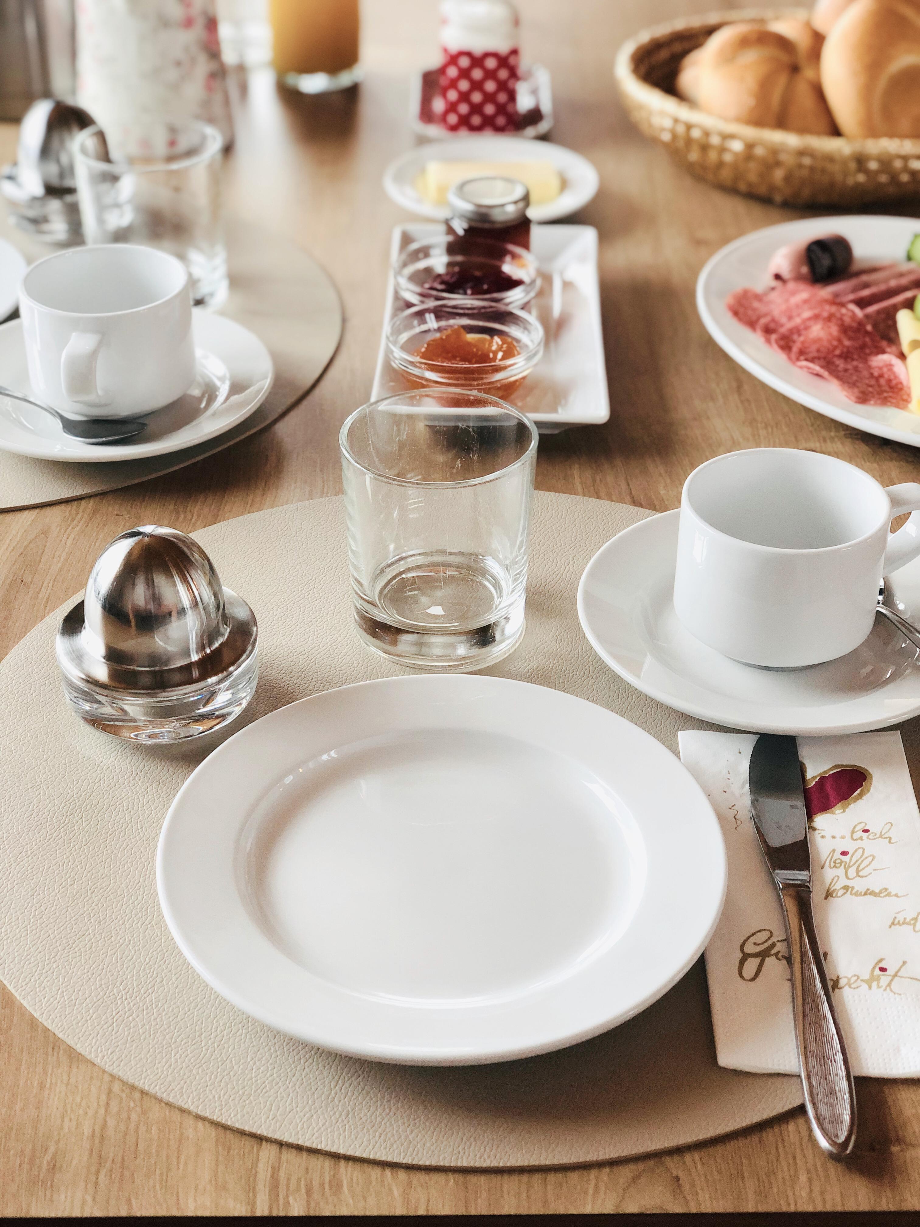 A beautifully set table for breakfast with plates, glasses, and cups. The table also features jam, butter, and fresh rolls.