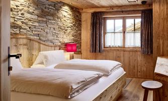 Naturally Sleep – Your Alpine Bedroom for Comfort. This lovingly furnished bedroom combines cozy old wood, a rustic stone wall, and modern design into a harmonious retreat. The large double bed with high-quality bed linen, warm light accents, and the natural wood paneling create a cozy atmosphere – perfect for restful nights in the mountains.