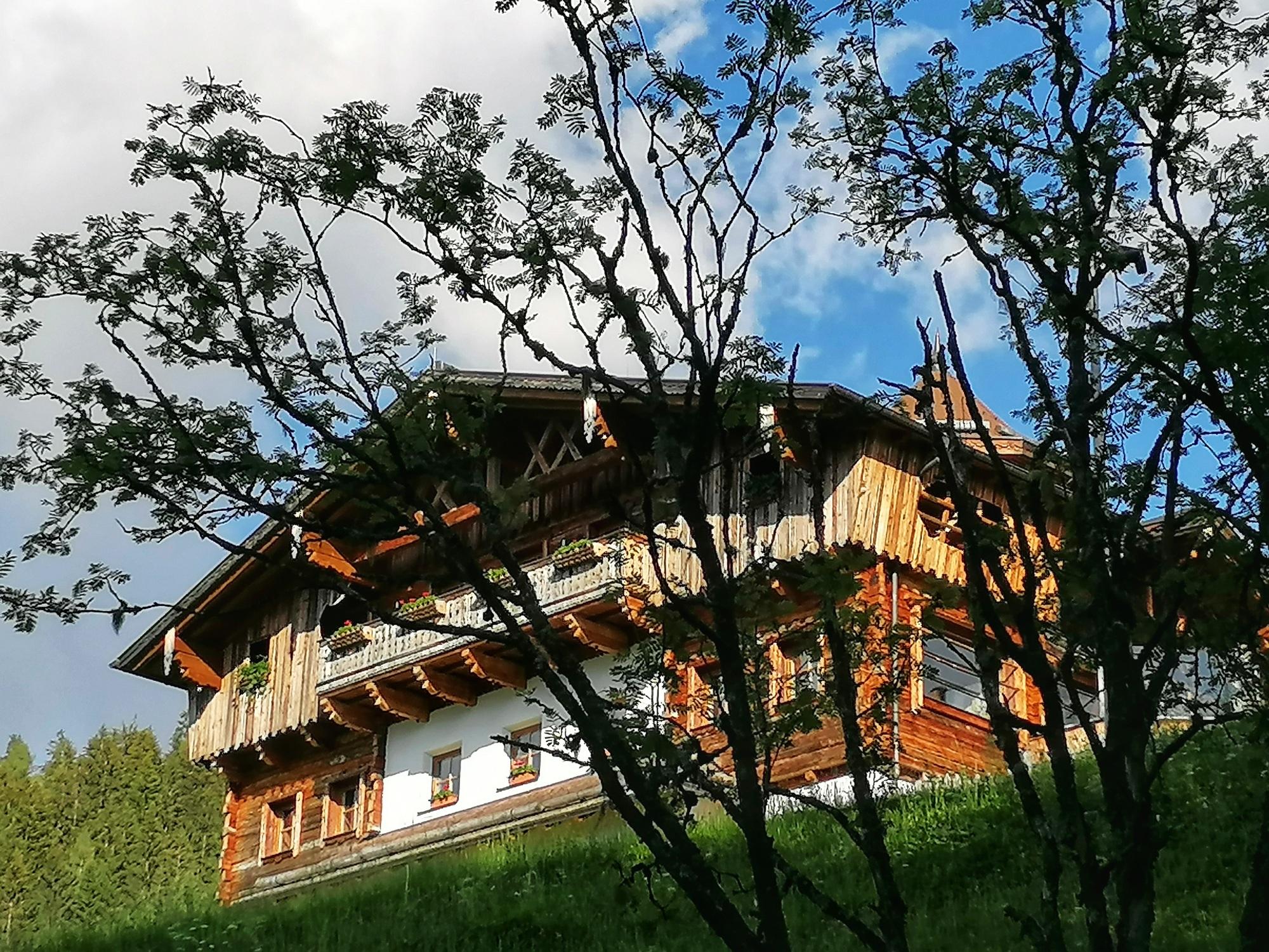 A traditional wooden house in the mountains, surrounded by trees. The sky is partly cloudy and it is green around the building.