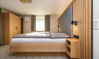 A modern bedroom with a double bed and wooden shelves. The room has large windows and a bright, friendly atmosphere.