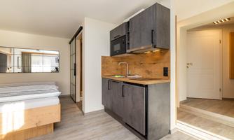 A modern kitchenette in a bright room with a cozy bed in the background. There are large windows that let in plenty of light.