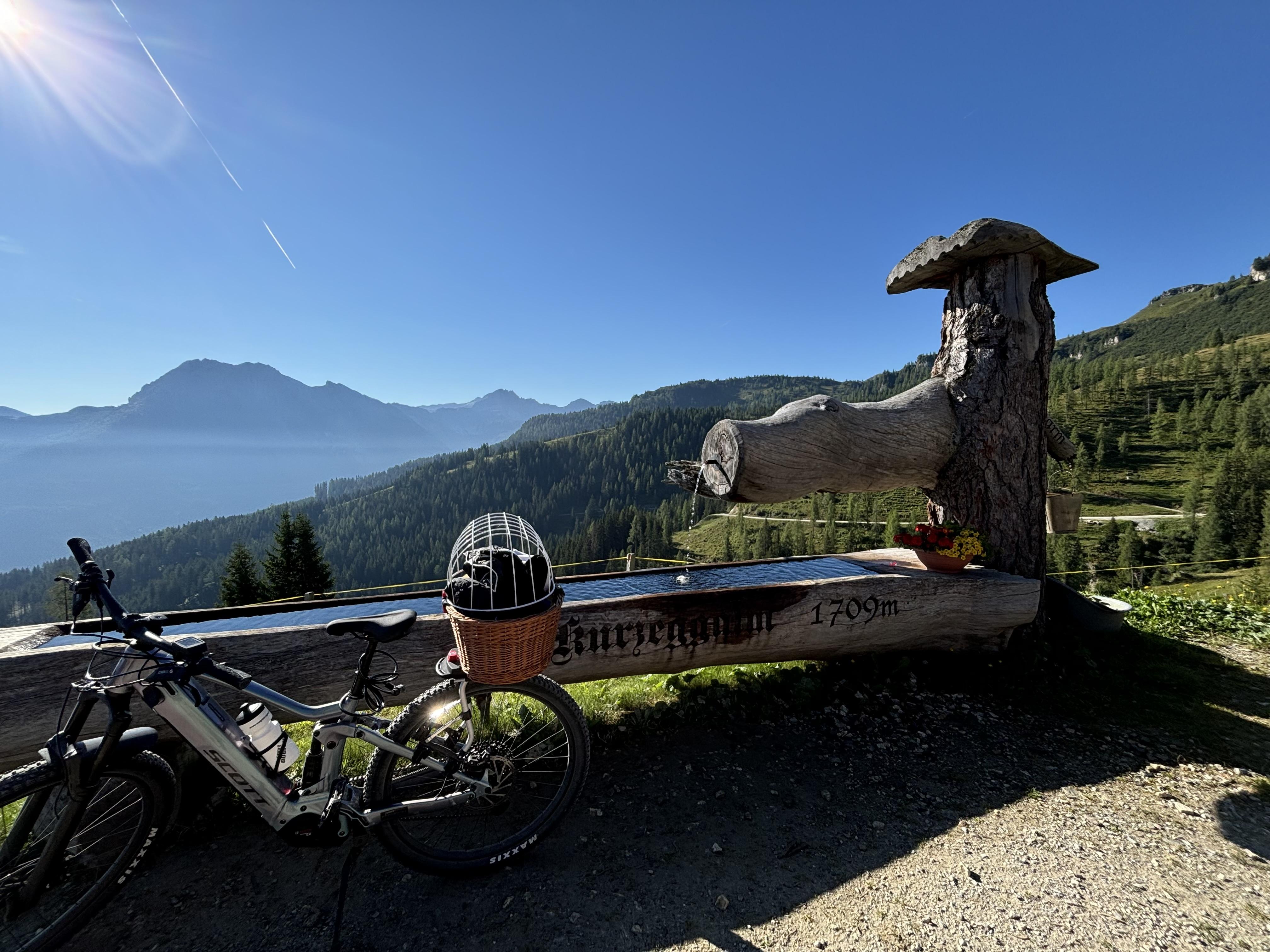 A picturesque mountain landscape with a wooden sign and a viewing platform at the Kurzegg-Alm. In the foreground stands an e-bike and a large fountain.