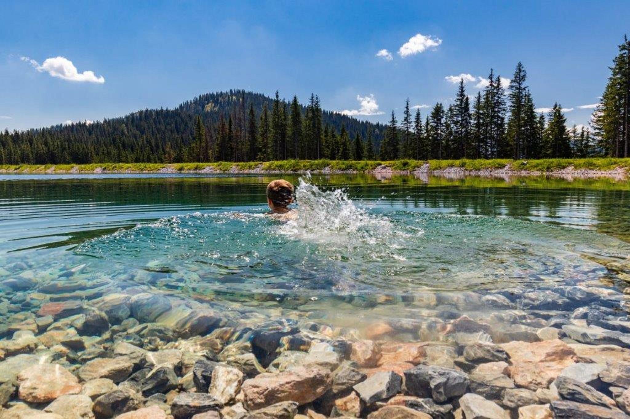 A clear lake surrounded by trees and mountains. A person is swimming in the water and creating splashes.