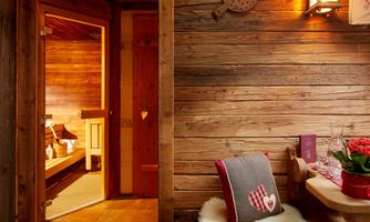 Private reclaimed wood sauna – relaxation in alpine style. Pure wellness in a cozy atmosphere: The stylish sauna made of fine reclaimed wood offers soothing warmth and relaxation after an active day in the mountains. The harmonious combination of natural materials, gentle lighting, and loving details creates a cozy retreat ambiance. Right next to it, a comfy relaxation area with fluffy sheepskin, heart decor, and fresh spring water invites you to unwind and catch your breath – the perfect place for body and soul.