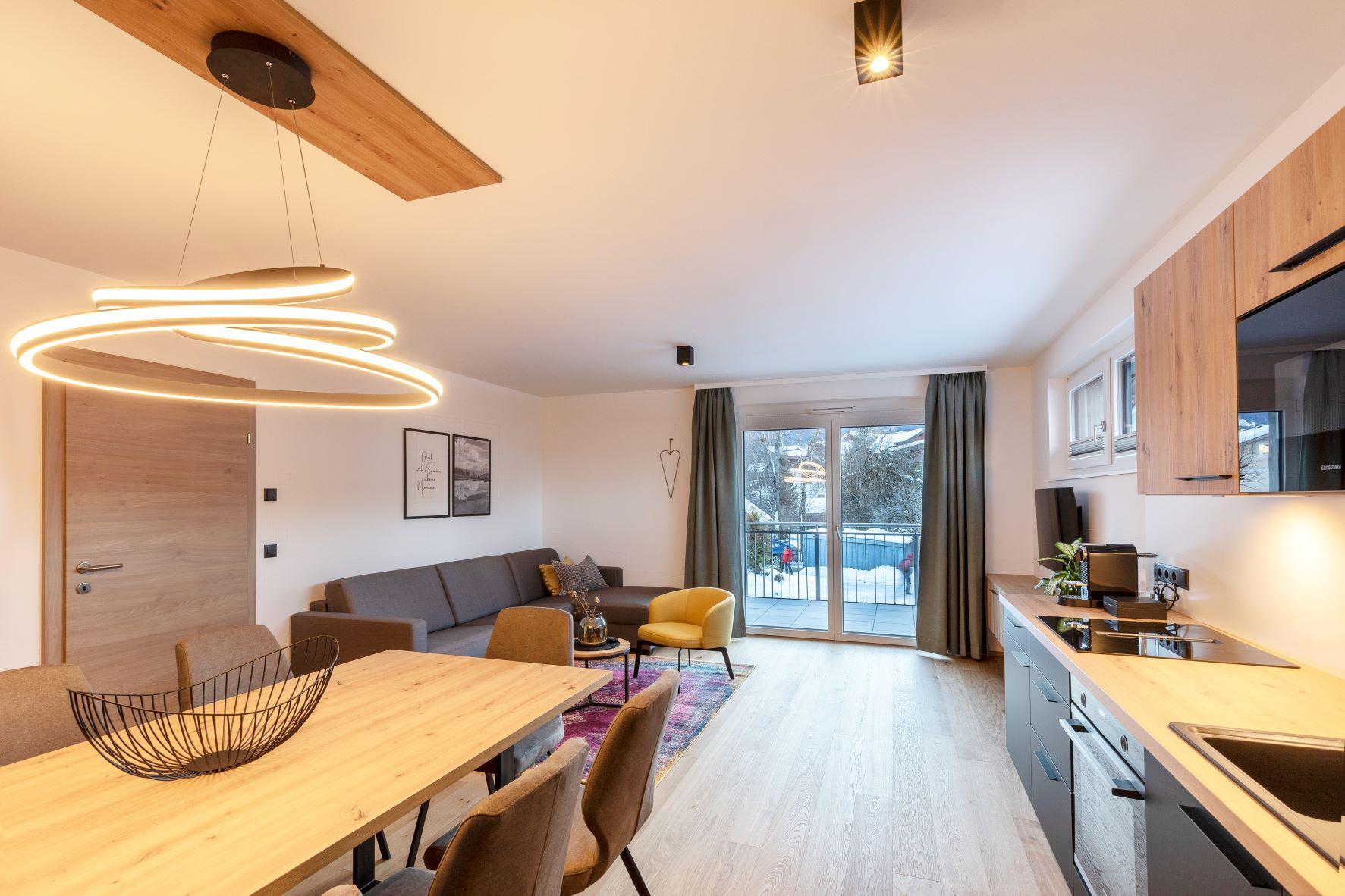 A modern living room with a dining table, a couch, and large windows. The room design is bright and friendly with wood and gray accents.