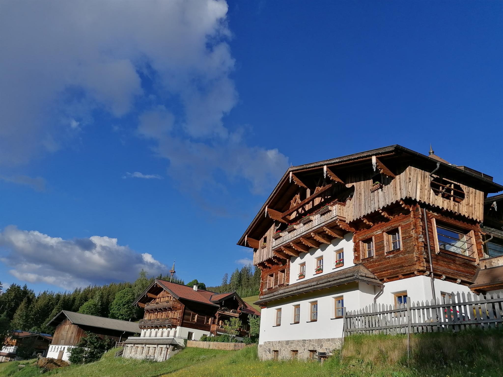 A house designed with great attention to detail, clad in reclaimed wood, nestled in the mountains and surrounded by green meadows. The sky is blue with a few clouds.
