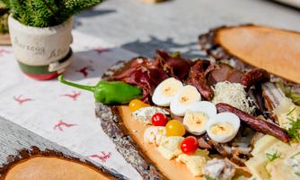 A rustic wooden board with various types of sausages and cheeses, decorated with boiled eggs and colorful tomatoes. In the background, there is a small pot with a plant.