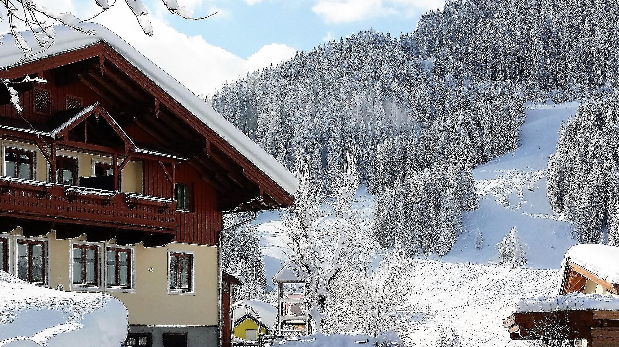 A picturesque alpine house in winter with snow-covered roofs and a snowy slope in the background. The trees are covered in snow and the sky is clear.