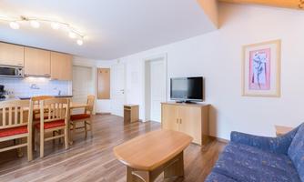A modern living and dining room with wooden floors and a kitchenette. The furnishings include a dining area, a sofa, and a television.