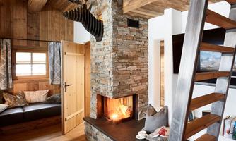 Living area with fireplace, cozy corner & alpine design. The stylish natural stone fireplace forms the centerpiece of this lovingly designed living space. With its three open sides, it provides comforting warmth and creates an inviting atmosphere. The cozy seating area with large cushions invites relaxation – perfect for reading hours or a glass of wine in the evening. Reclaimed wood, luxurious textiles, and modern details such as the ladder to the gallery level and an integrated Sonos system combine alpine charm with contemporary living comfort.