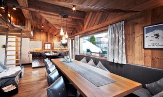 Generous living and dining area with panoramic window. The open living space captivates with cozy cabin flair and modern comfort. The combination of natural wood, stylish designer lights, and high-quality furniture creates a warm, inviting atmosphere. The large dining table with surrounding bench and comfortable chairs offers space for social gatherings. A special highlight is the large panoramic window, which not only lets in plenty of light but also provides a view of the mountain landscape – a place to arrive and enjoy.