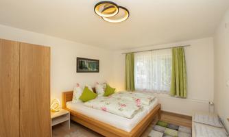 A cozy bedroom with a large bed and colorful pillows. There is a wooden wardrobe and bright curtains that let the light in.