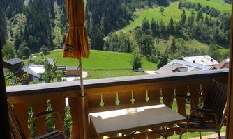 A balcony with two chairs and a table, surrounded by green hills and forest. In the background, there are some houses and a bright blue sky visible.